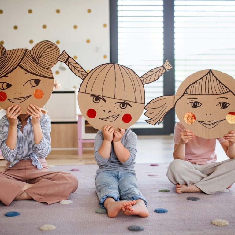 Drei Kinder sitzen auf dem Boden und halten große, bemalte Pappmasken vor ihren Gesichtern.